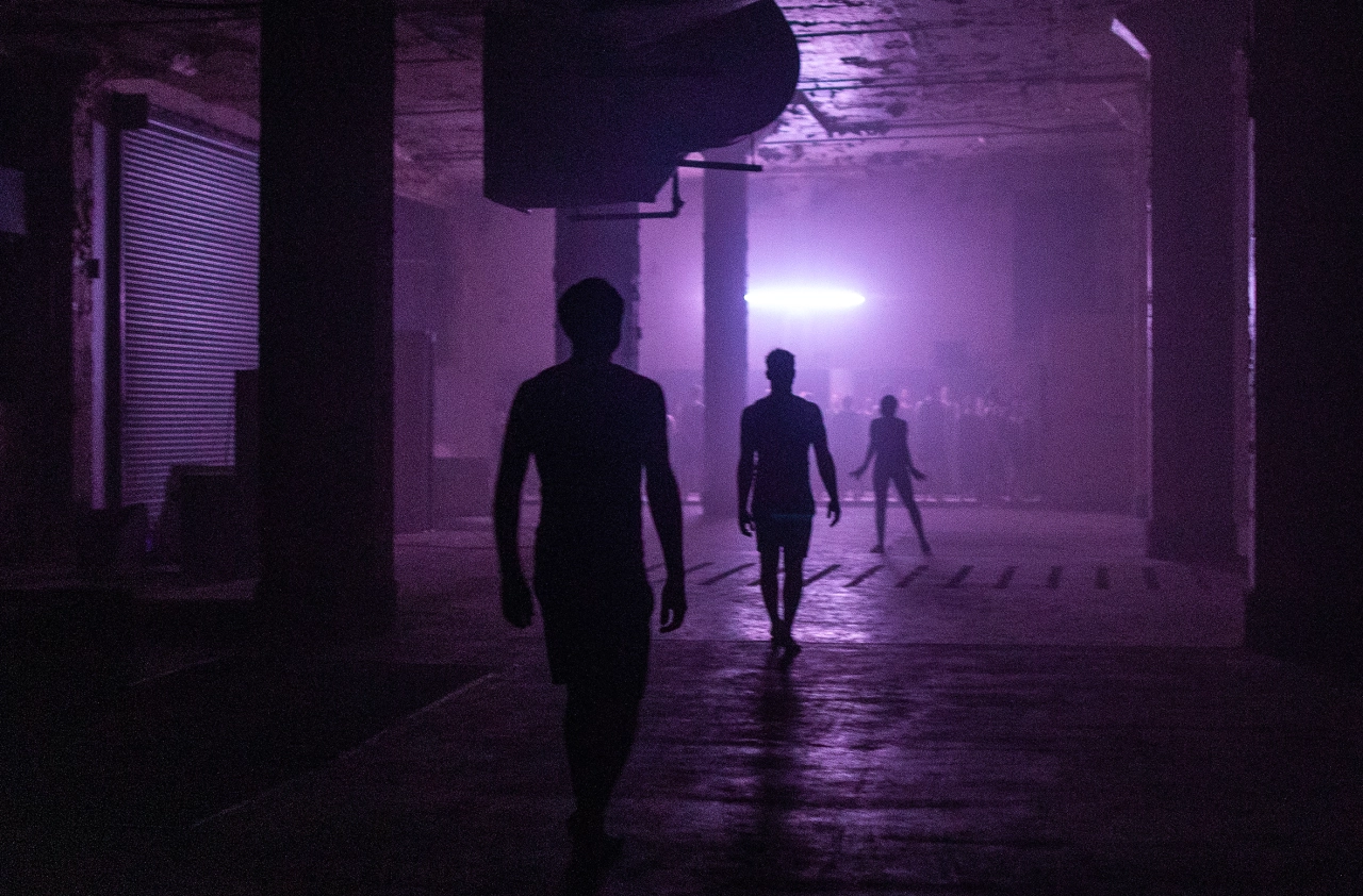 Silhouetted figures move through a dimly lit warehouse space bathed in purple light, with industrial columns and a suspended light fixture visible overhead