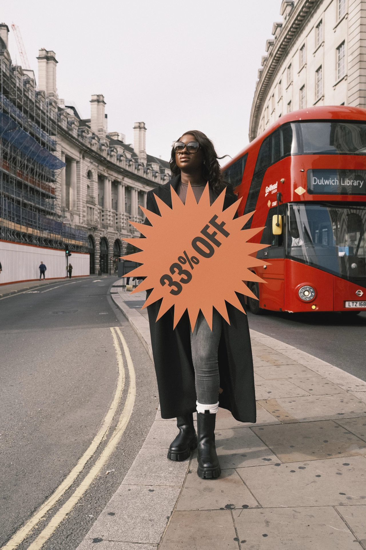 Woman in black coat and sunglasses holding an orange starburst graphic reading "33% off" on a London street beside a red double-decker bus
