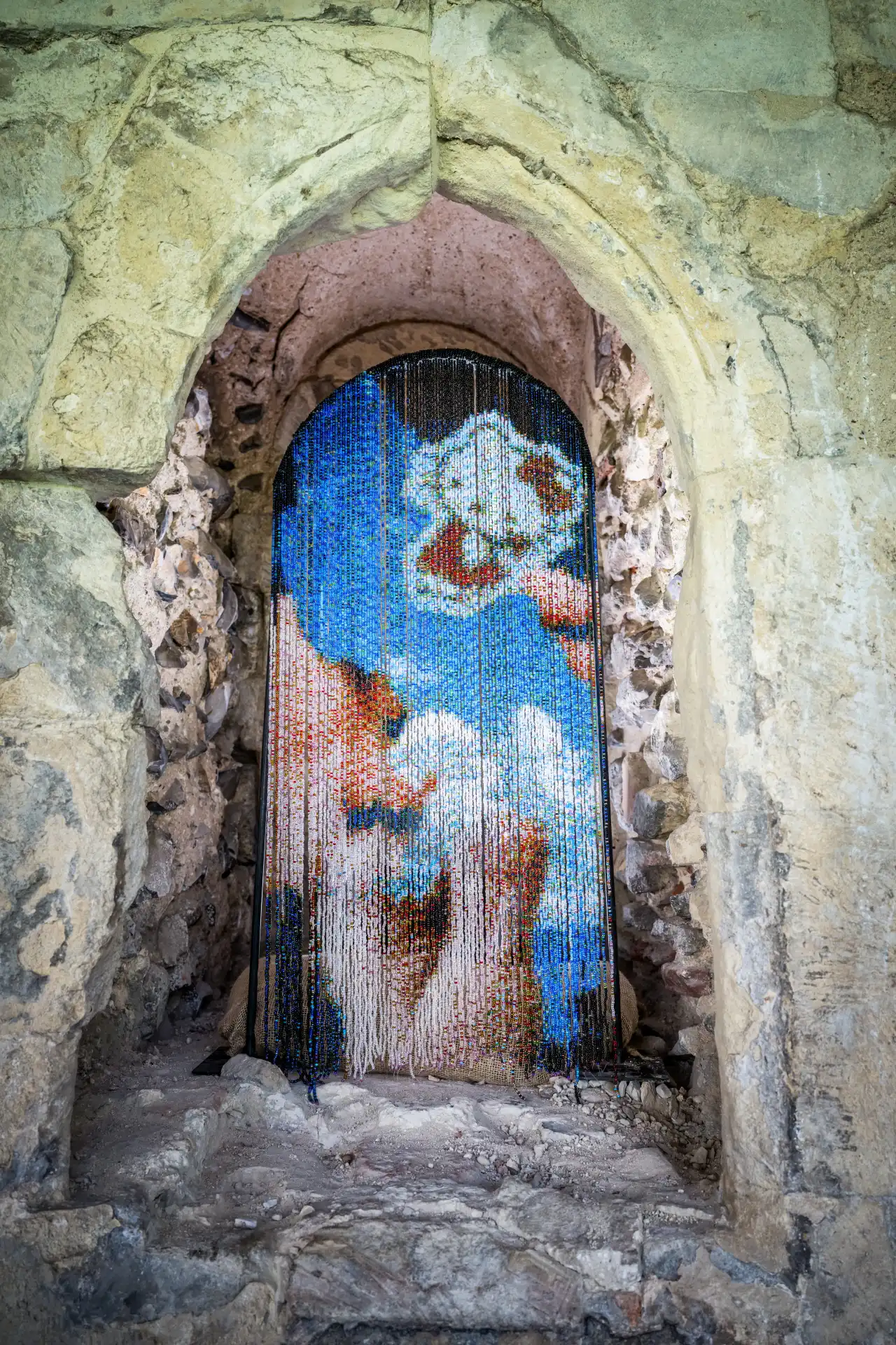 Archway in stone wall frames a narrow doorway with beaded curtain depicting blue sky and white clouds with rust-coloured marks, by Lily Bunney