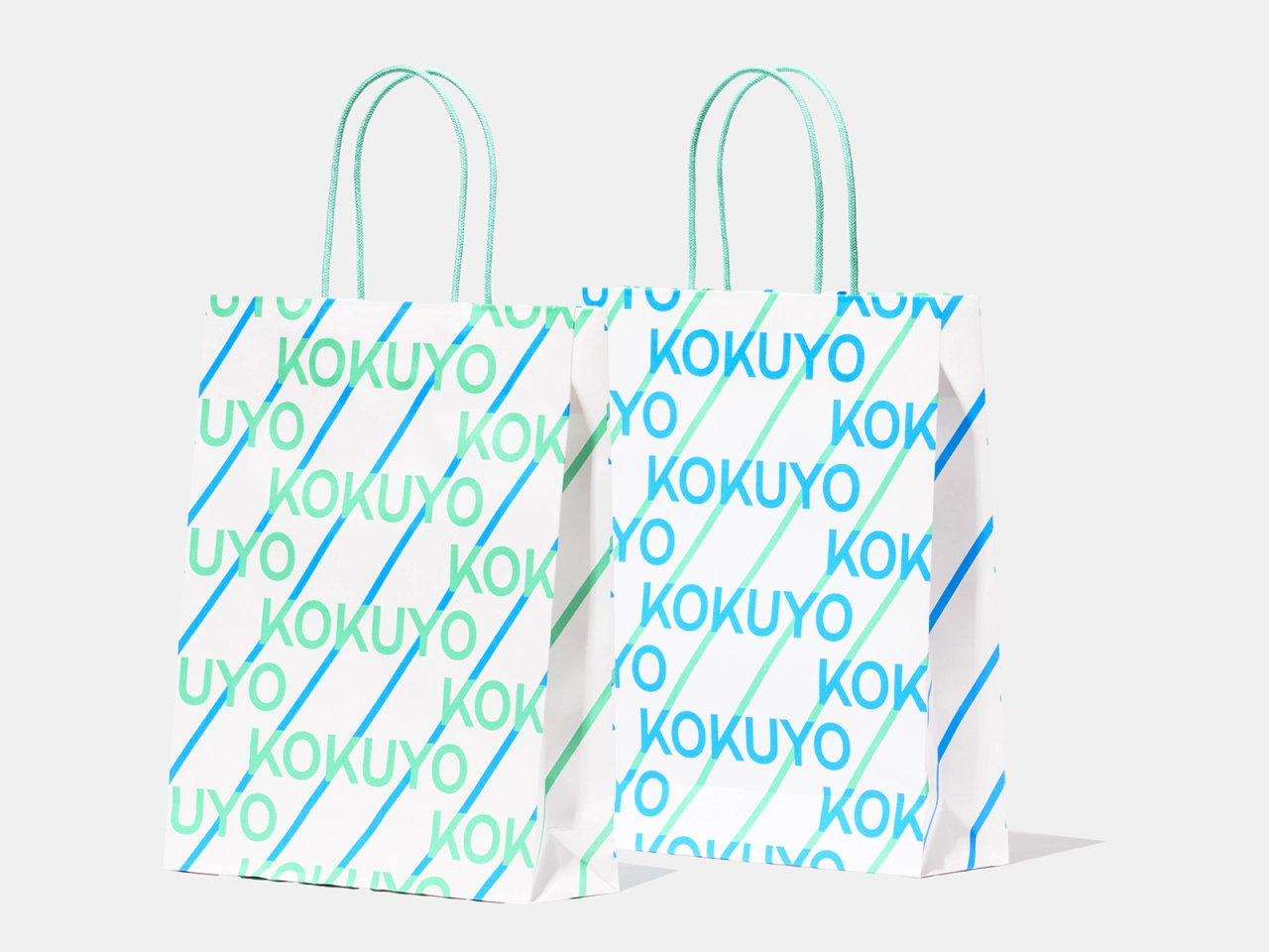 White carrier bag with diagonal repeating "KOKUYO" text in gradient shades of blue and green, designed by Kanaisasaki