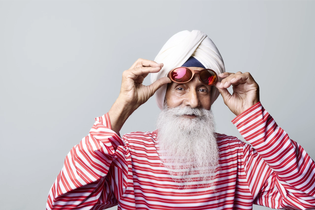 Man with white beard and hair wearing red and white striped shirt and white cap adjusts pink-tinted goggles while looking at camera against grey background