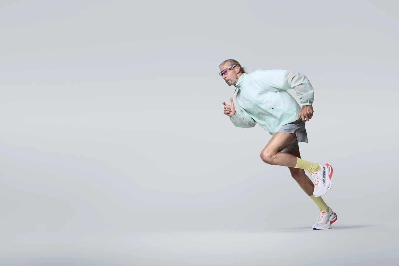 Older man wearing glasses and light blue jacket runs energetically against a white background, wearing yellow shorts and trainers