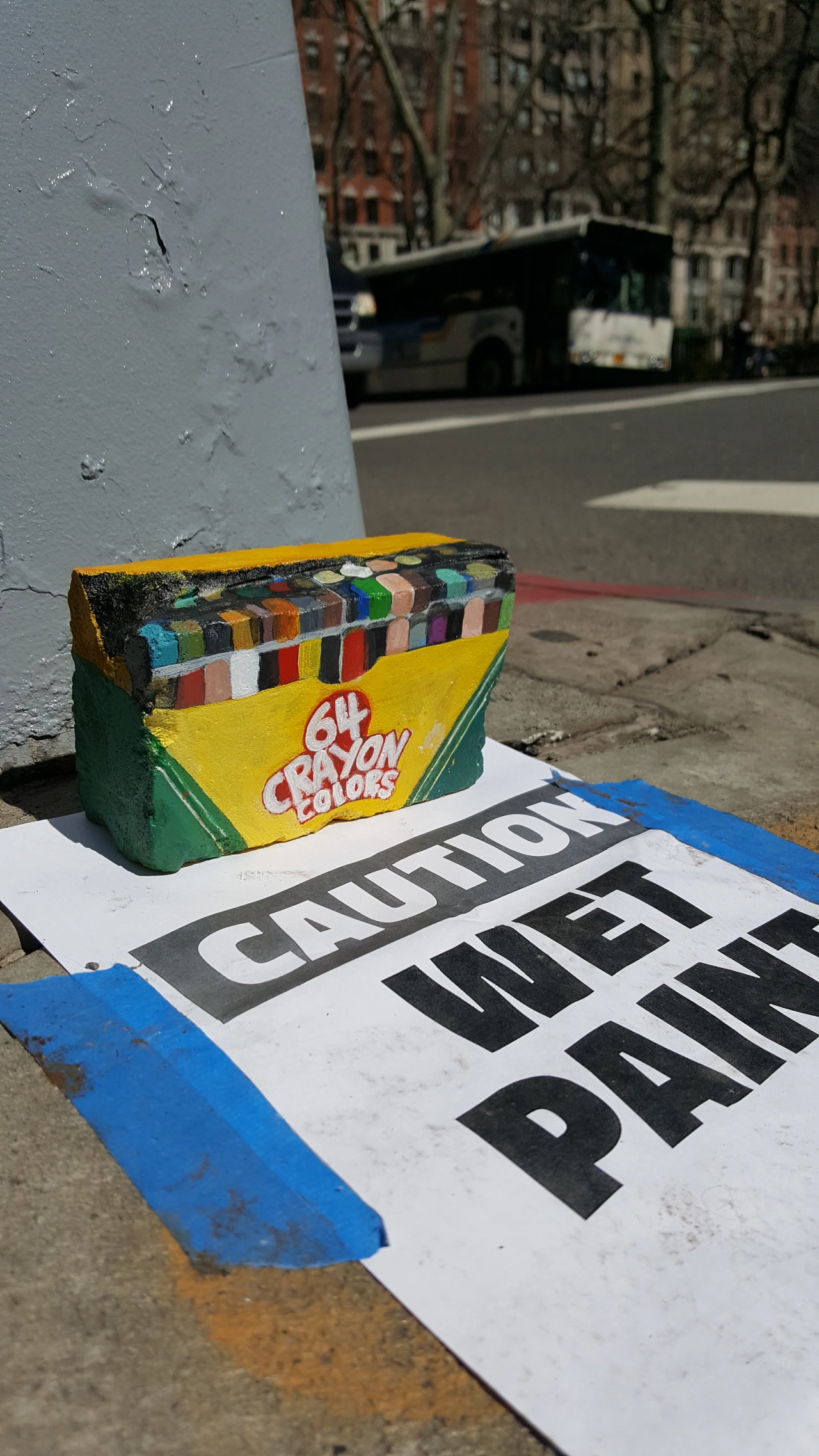 Colourful Crayola box painted on a rock beside a white sign reading "Caution Wet Paint" on a city street