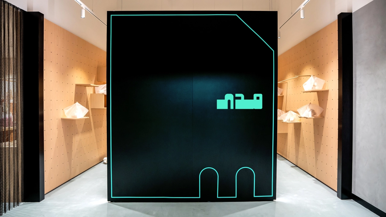 Neon green pixelated text and geometric shapes displayed on a black screen with cyan border, installed in a minimalist corridor with warm-lit alcoves on either side