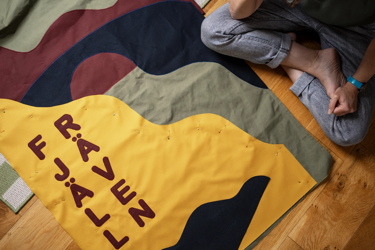 Person sitting beside hand-stitched fabric banners by Miscellaneous Adventures for Fjällräven, with yellow banner displaying text in burgundy lettering