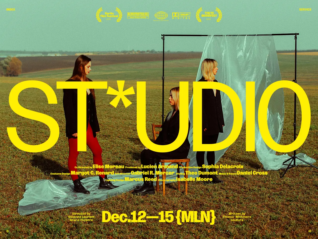 Three people stand in a field beside a transparent plastic sheet suspended on a metal frame, with festival laurel wreaths and dates displayed above in yellow text.