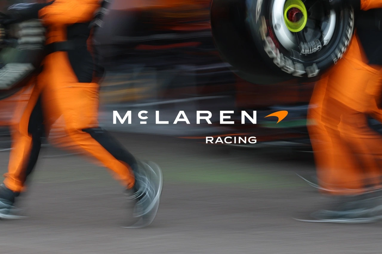 McLaren Racing team members in orange uniforms working on a Formula 1 car during a pit stop, with the new McLaren Racing wordmark and logo displayed