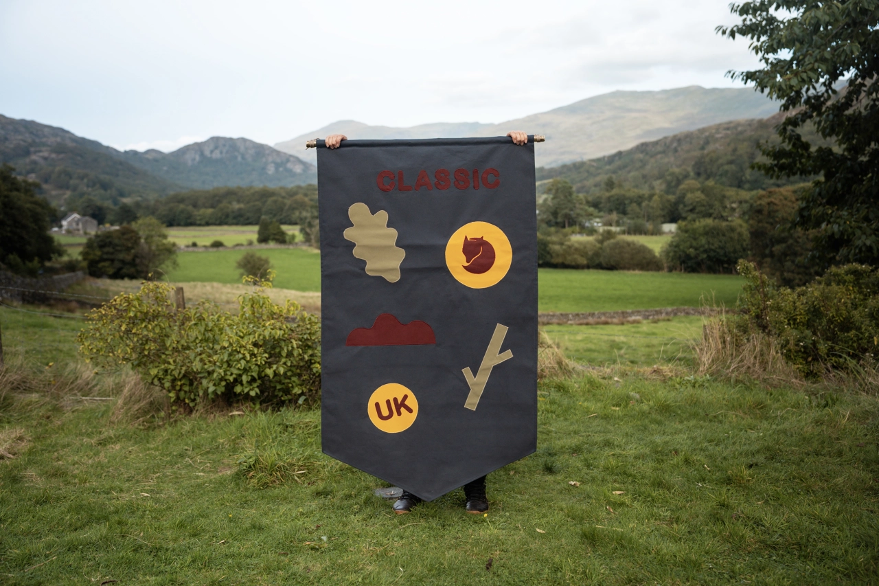 Large dark fabric banner hand-stitched with yellow and red symbols including a circle, oak leaf and UK badge, displayed in a Lake District valley landscape