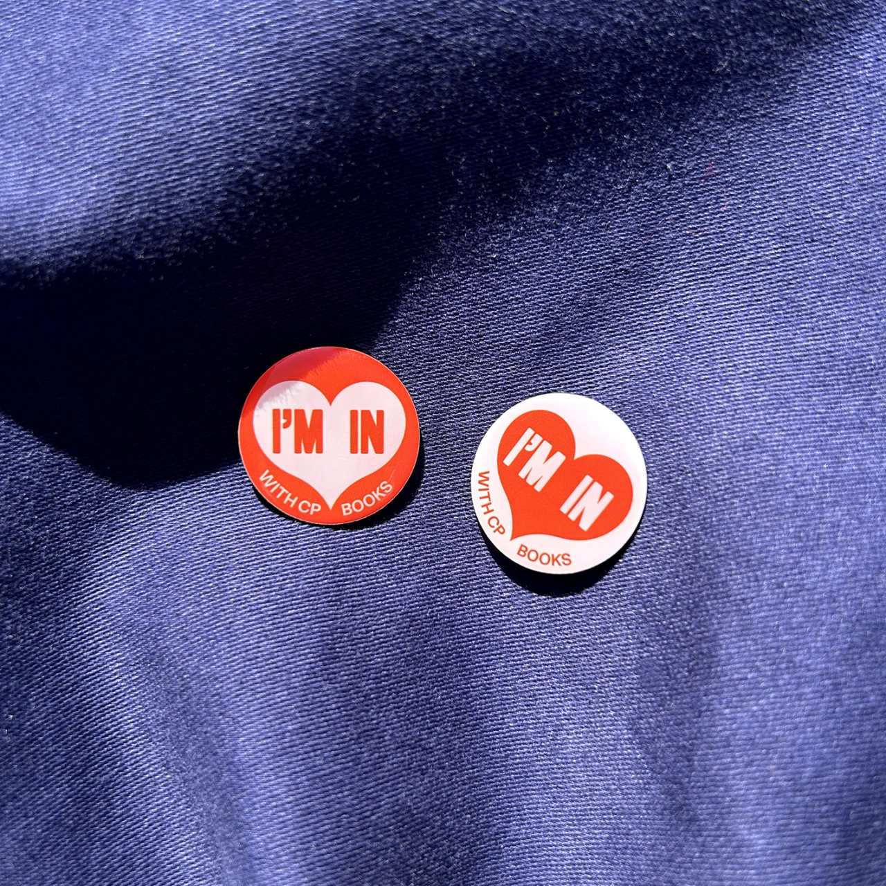 Two circular pin badges on blue denim, one red with "I'M IN" text and one white with "I'M IN" text, both branded with Witchop Books logo