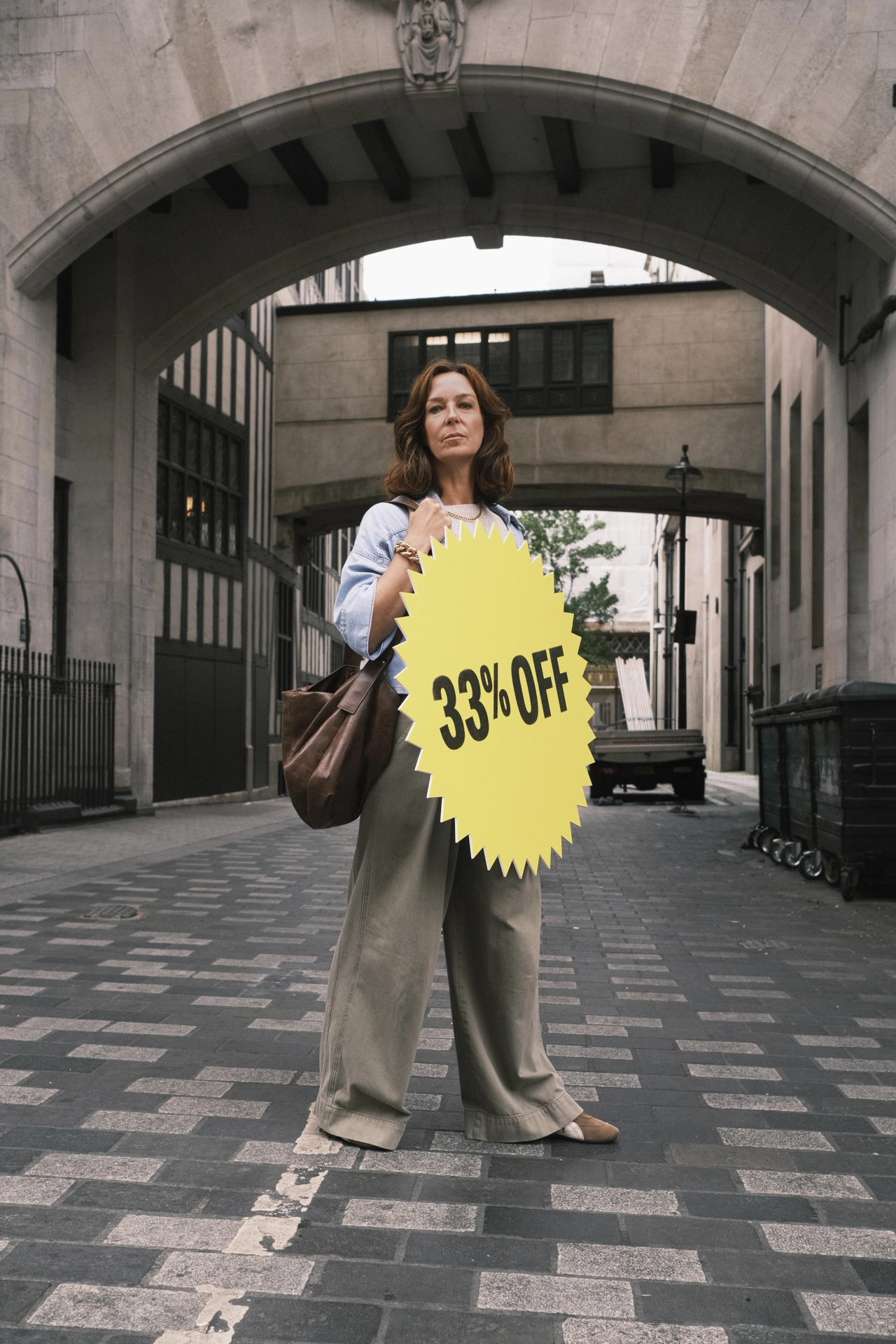 Woman holding a bright yellow starburst sale sign reading "33% OFF" in front of her face whilst standing in a stone archway