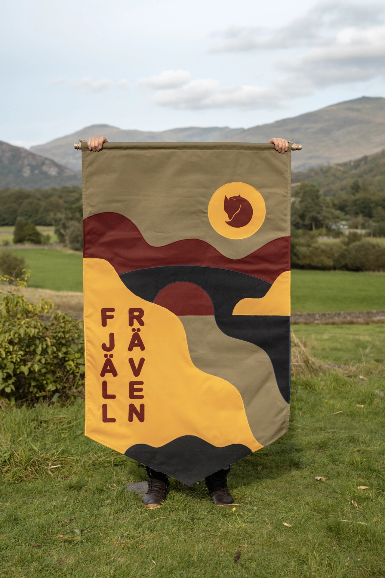 Person holding a large hand-stitched fabric banner featuring geometric mountains, yellow sun and moon, and "Fjäll Råven" text, standing in a Lake District valley with hills behind