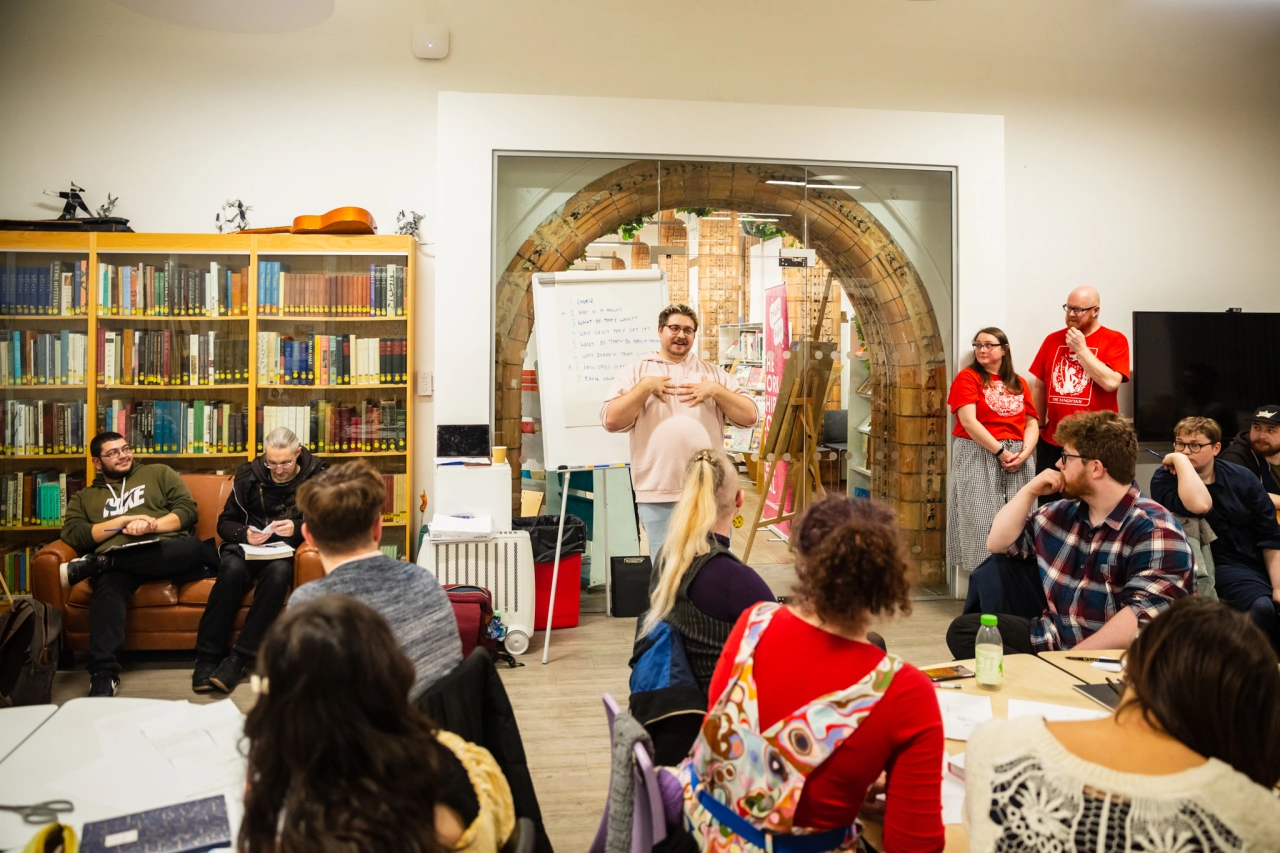 A presenter gestures while speaking to a seated audience in a community space with bookshelves, while attendees in red Thought Bubble t-shirts stand nearby