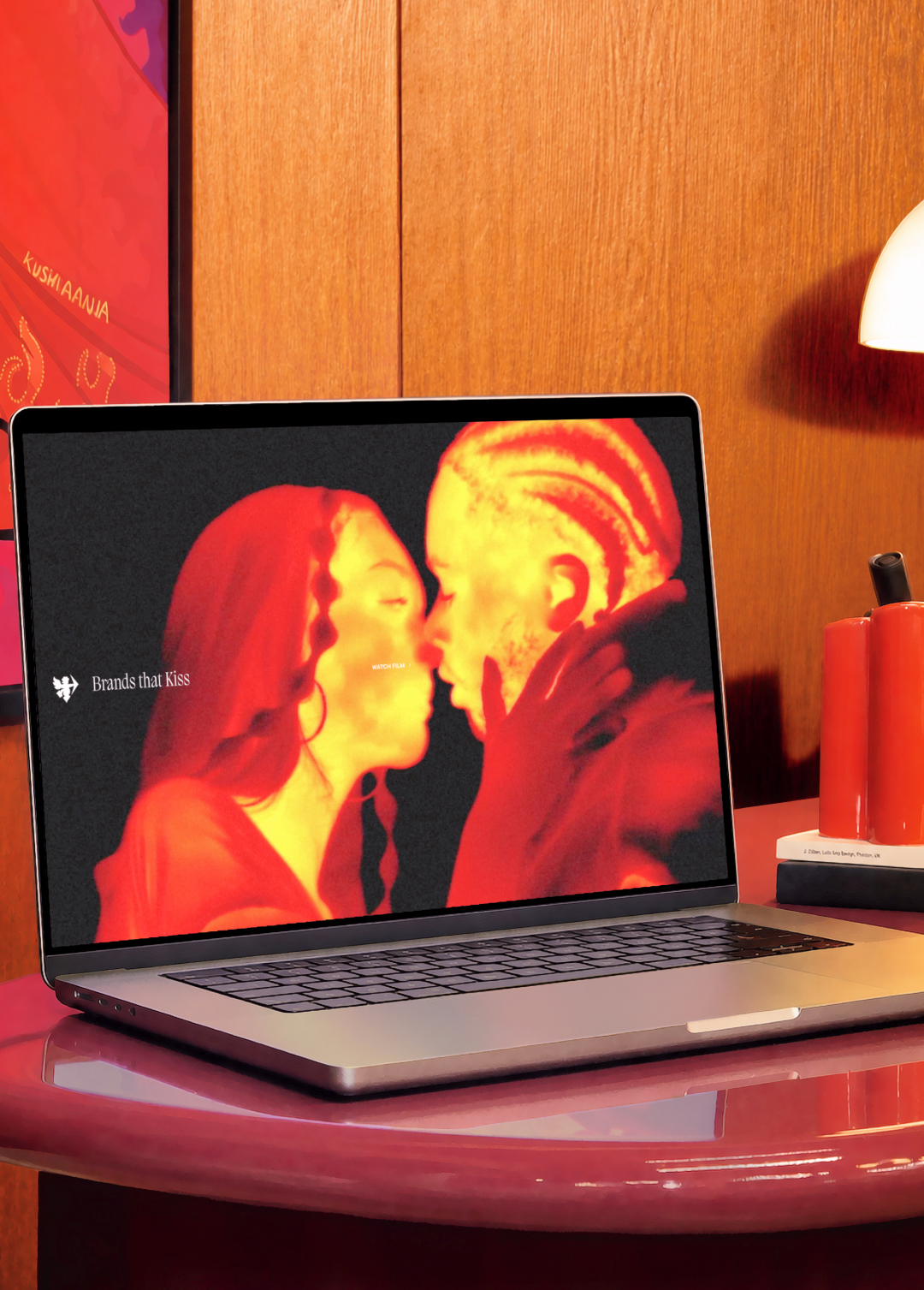 Laptop displaying a thermal-style image of two people about to kiss in red and yellow tones, positioned on a red desk with wooden panelling behind it