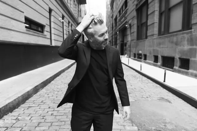 Man in black suit standing in an urban alleyway, hand touching his head, looking to the side in black and white photography