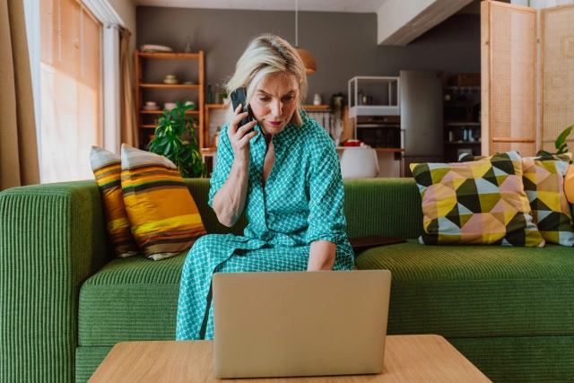 Woman in turquoise pyjamas sitting on green sofa with laptop, speaking on phone in bright, plant-filled living room