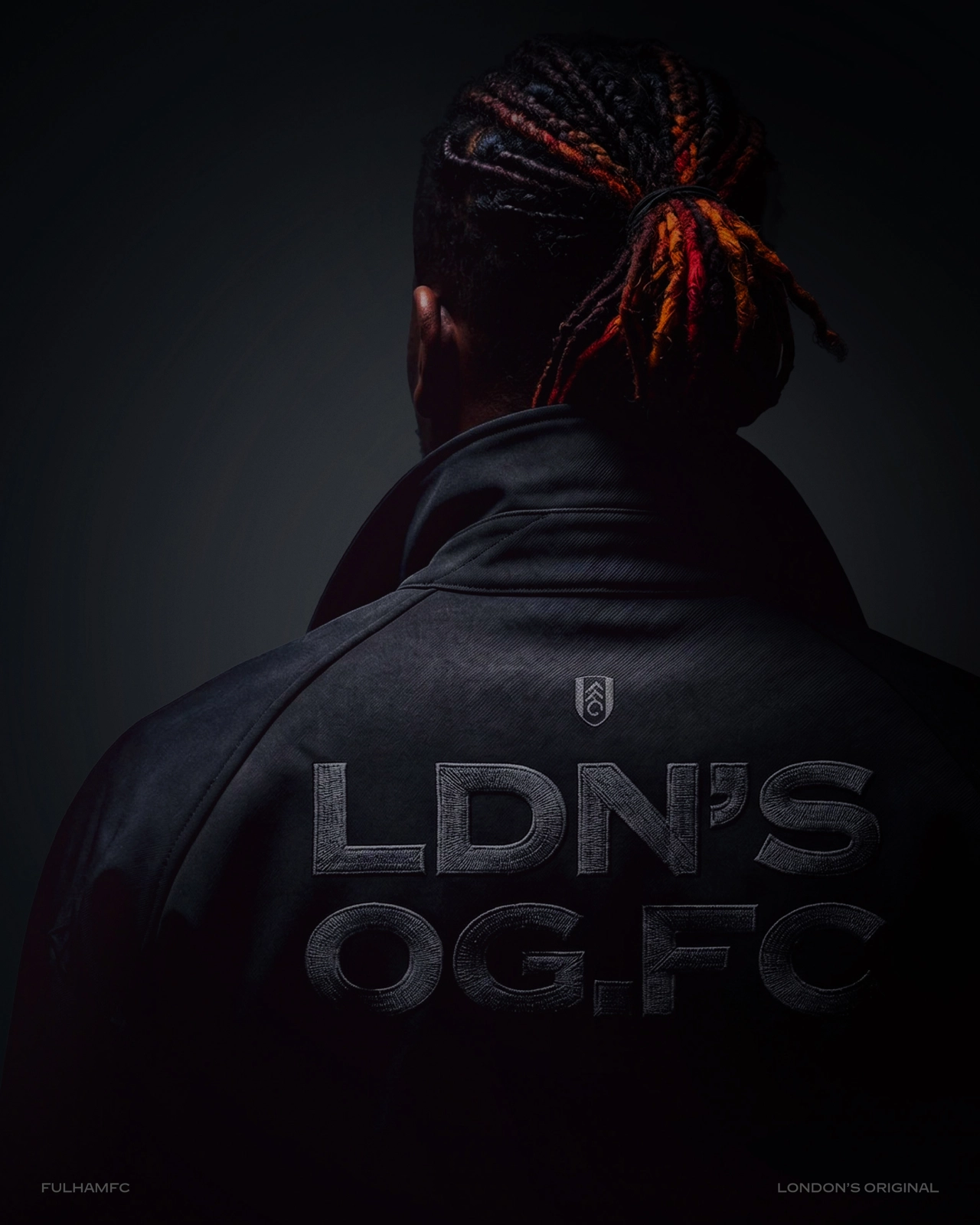 Person wearing a black Fulham FC jacket with red dreadlocks tied back, photographed from behind against a dark background