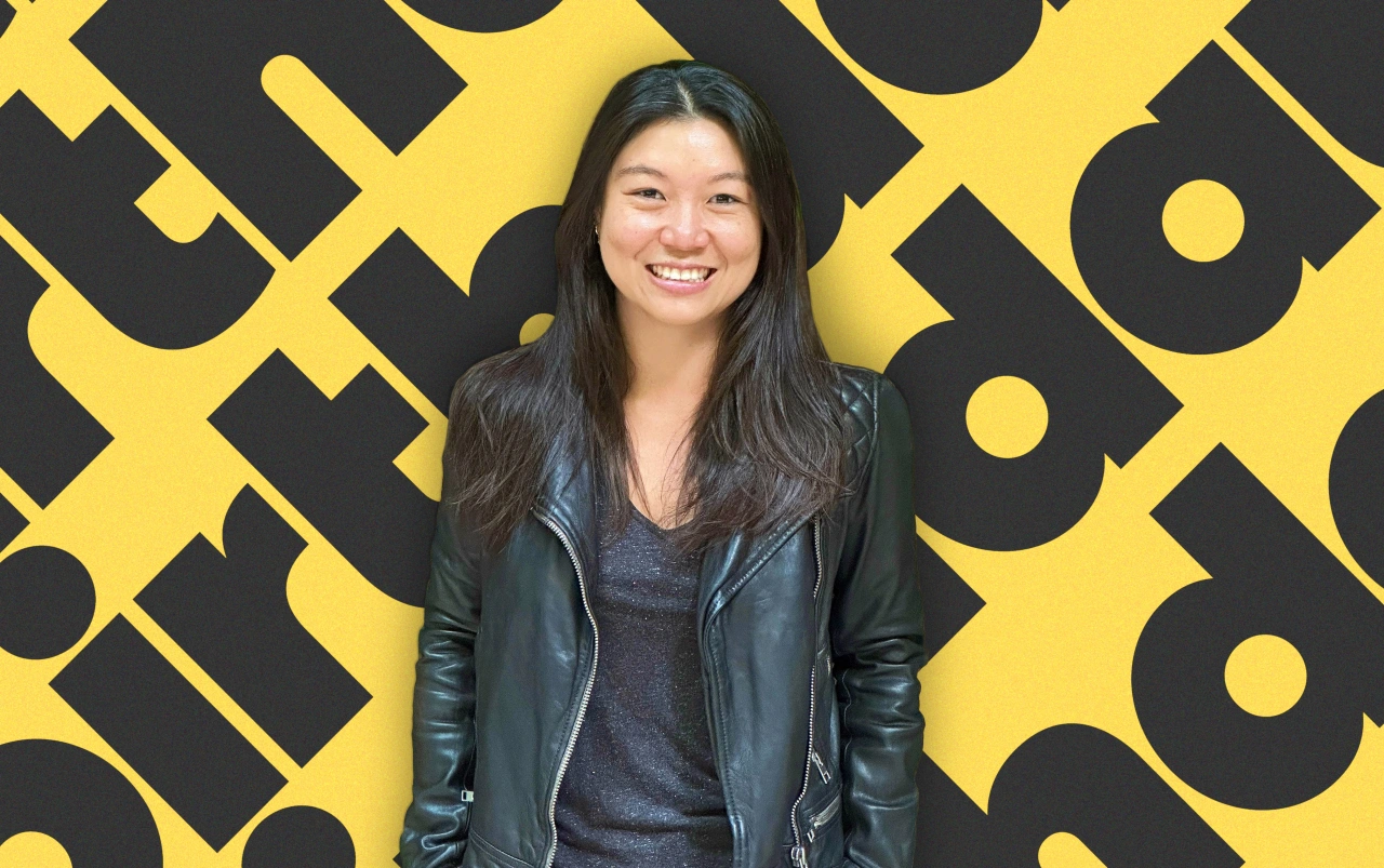 Woman with long dark hair smiling at camera, wearing black leather jacket over grey top, standing in front of yellow and black geometric patterned background