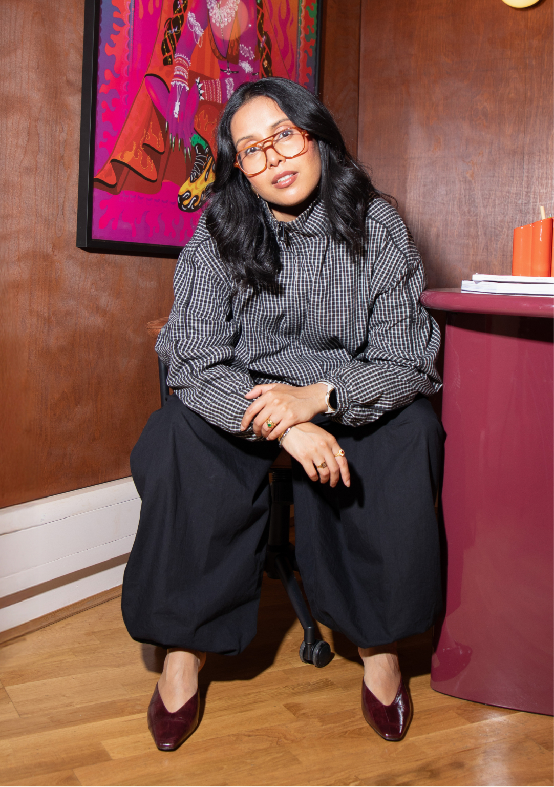 Poonam Saini Kilb sits in a studio space wearing glasses and a grey checked shirt, with colourful artwork visible on the wall behind her