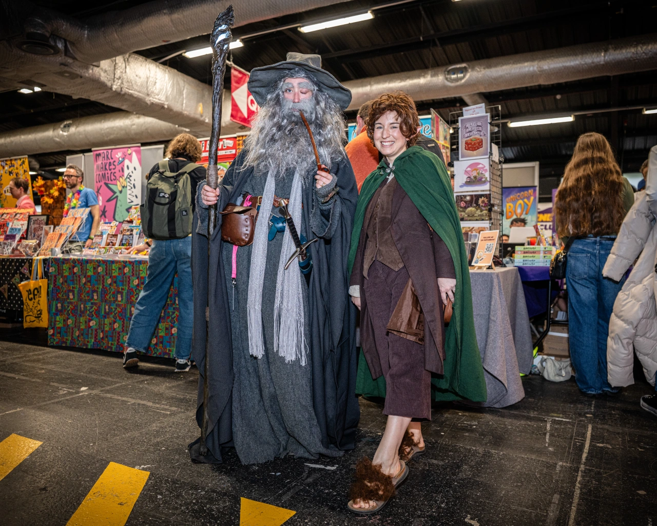 Cosplayers dressed as Gandalf and Bilbo Baggins from The Lord of the Rings at Thought Bubble comic art festival, surrounded by vendor stalls