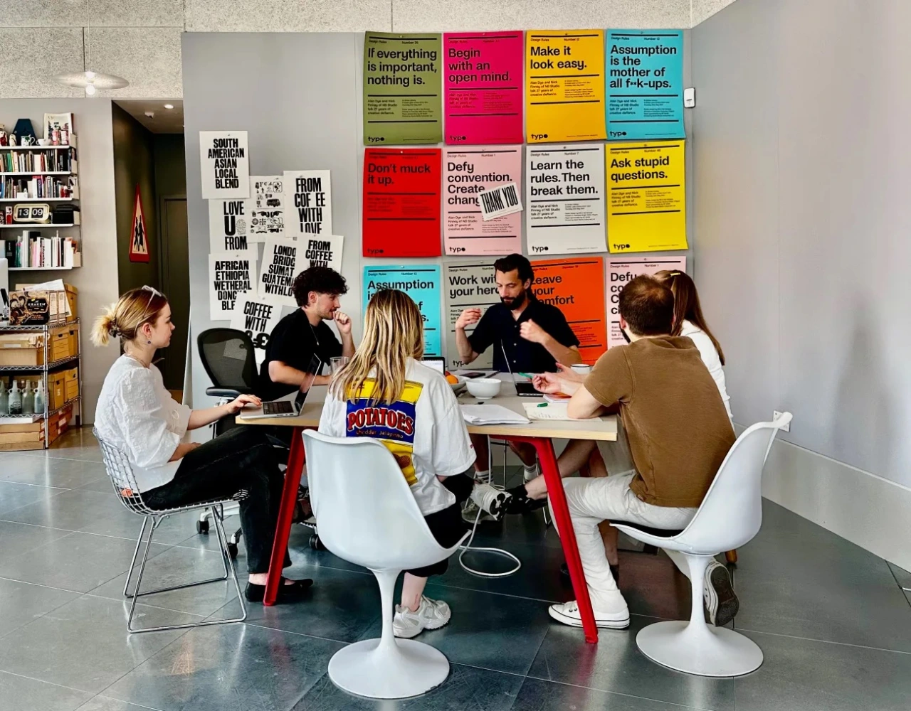 Five people sit around a red and white table in a studio space with colourful typographic posters covering the wall behind them.