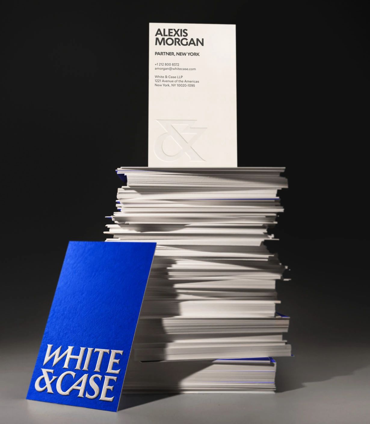 Alexis Morgan's business card for White & Case positioned on top of a tall stack of white papers, with the firm's blue branded folder leaning against it