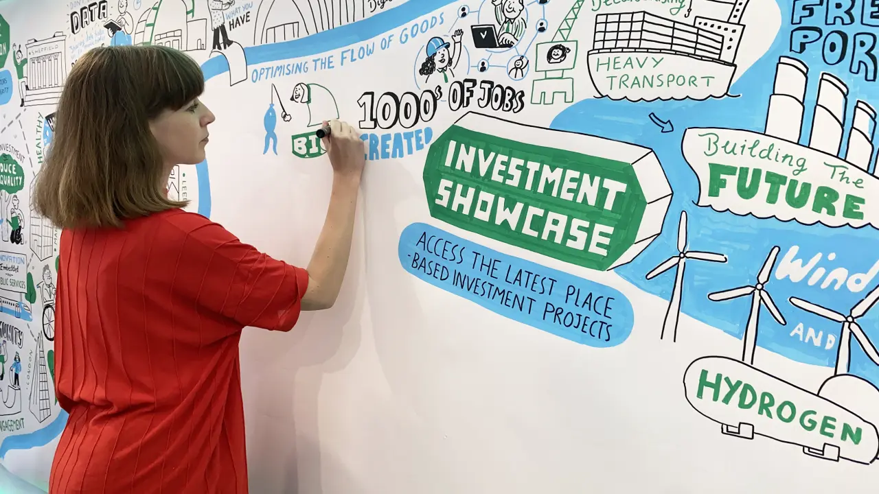 Woman in red shirt drawing on a large mural featuring investment showcase graphics and renewable energy illustrations including wind turbines and hydrogen power symbols