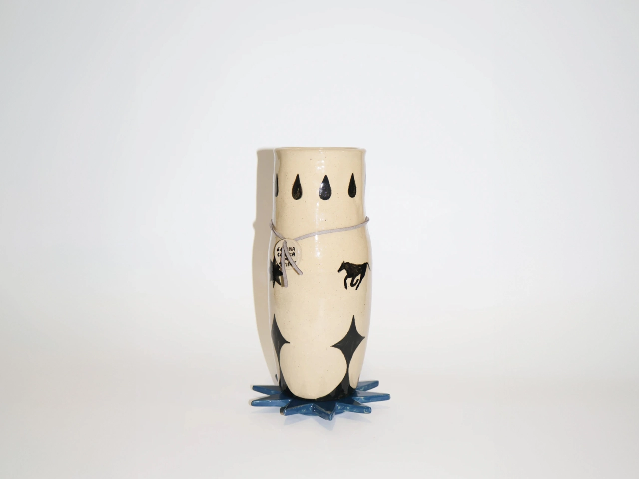 Cream-coloured cylindrical ceramic vessel with black drawn details including raindrops, a horse and geometric shapes, mounted on a blue base