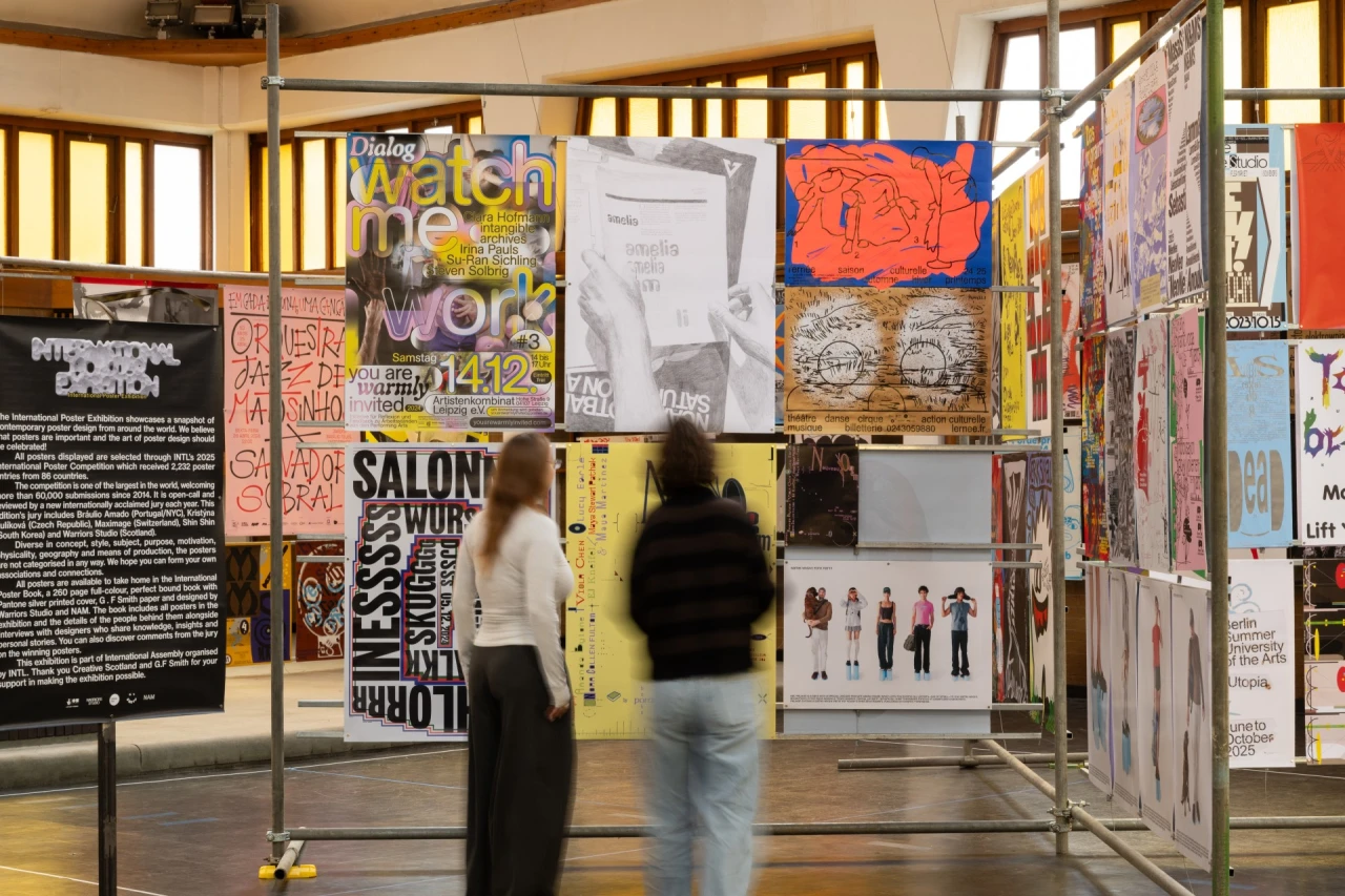 Two visitors walk through an exhibition space displaying colourful posters and design work on panels and walls, part of the International Poster Exhibition
