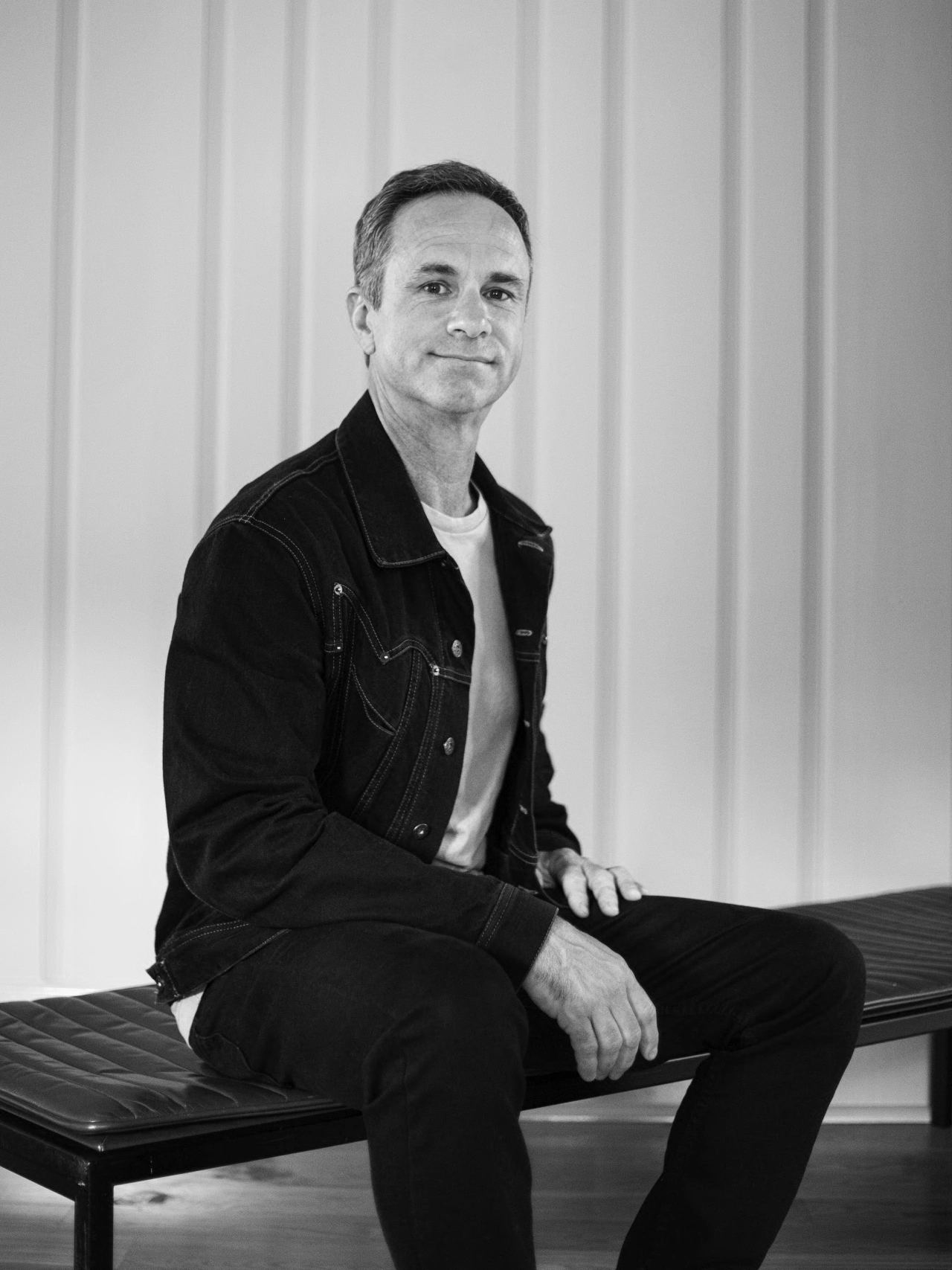 Man in black denim jacket and white shirt sitting on a bench against a white striped background