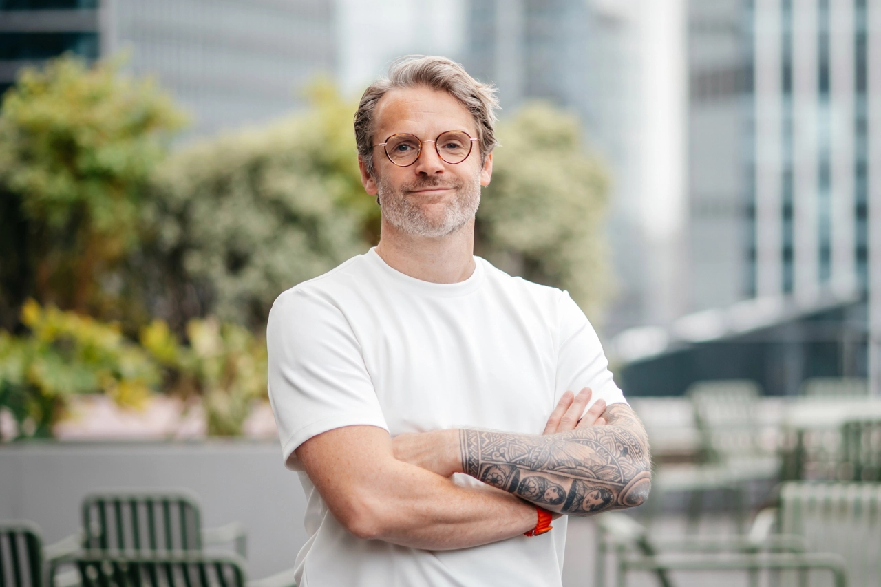 Man with short blonde hair and glasses wearing a white t-shirt, standing with arms crossed in front of a blurred urban rooftop setting