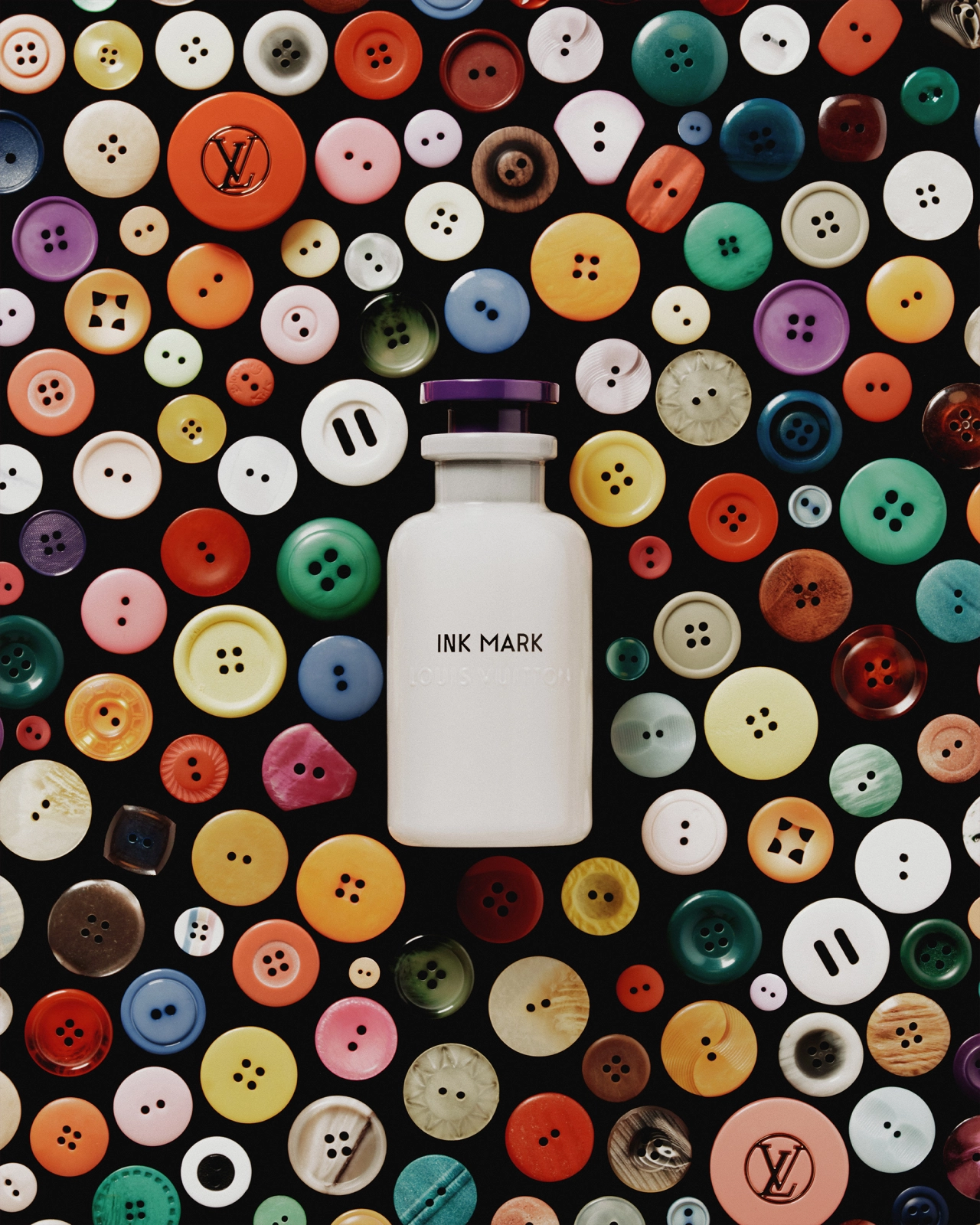 White bottle labelled Ink Mark surrounded by multicoloured sewing buttons in various sizes on black background