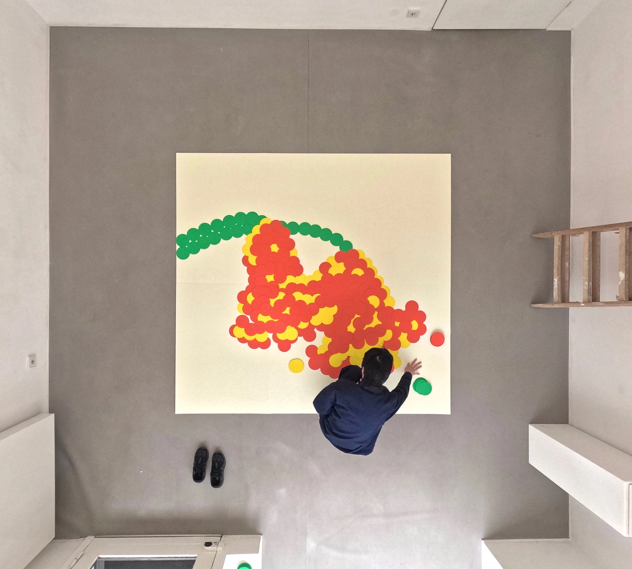 Ayong Son reaches up to place a large hand-cut red and yellow paper flower onto a cream-coloured canvas mounted on a grey wall, with green leaves arching above it