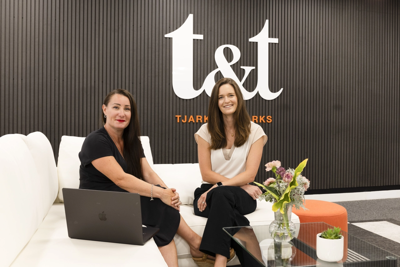 Donna West and Alex Poulton sit on a white sofa in front of a dark corrugated wall displaying the Tjarks & Tjarks logo, with a laptop and flowers on the table beside them