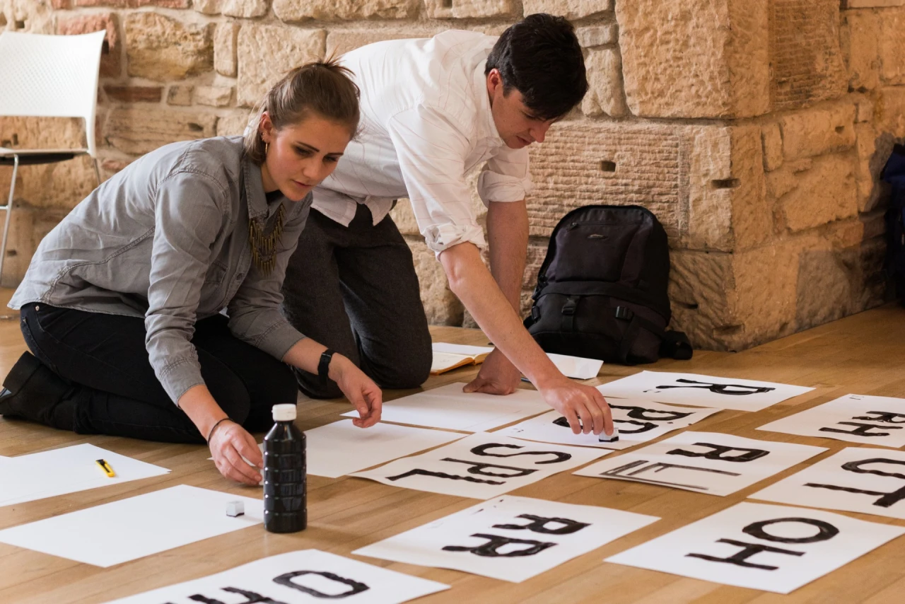 Two people kneeling on a wooden floor examine sheets of large printed letters and text layouts arranged in a grid pattern against a stone wall.