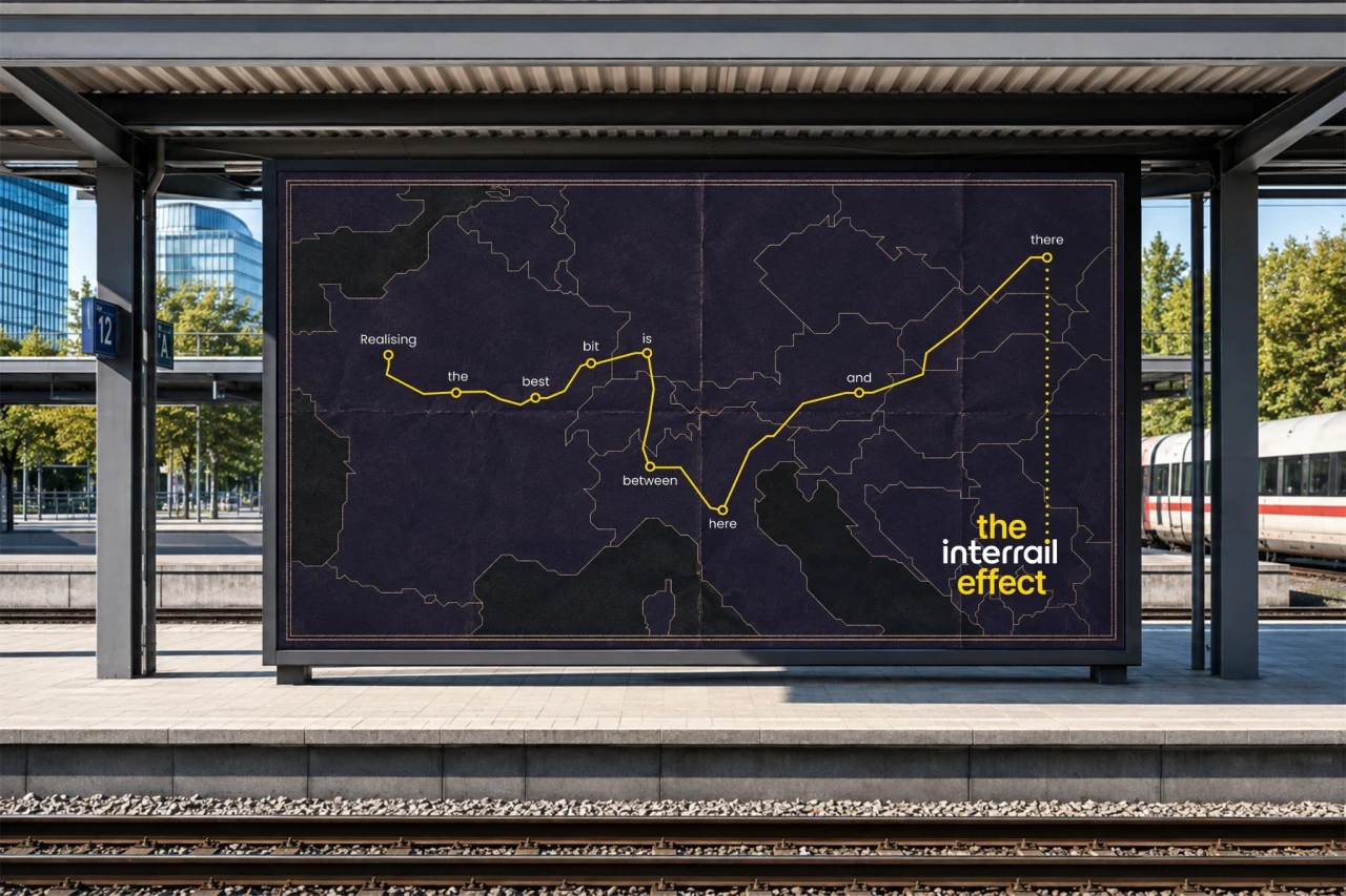 Interrail advertisement displayed on a station shelter showing a yellow train route with text labels forming the phrase "Realising the best bit is between here and there and there" overlaid on a map of Europe for Interrail campaign