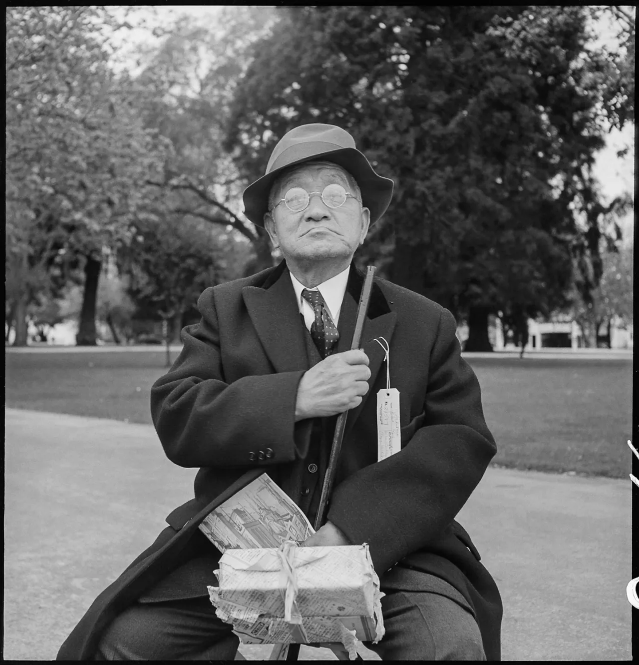 Man in suit and hat holds a walking stick and bundle of possessions while sitting on a bench in a park, looking upward with a grave expression