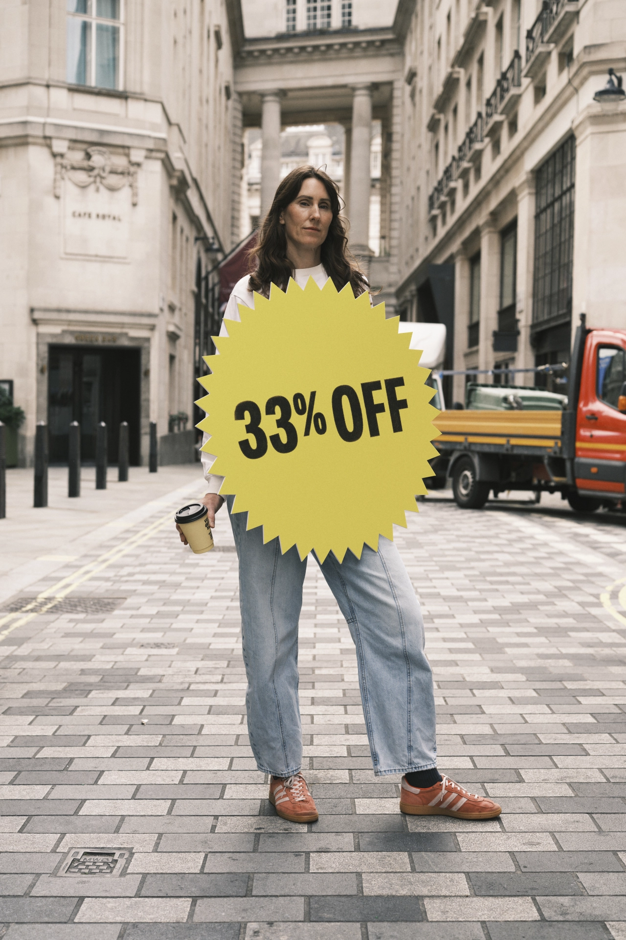 Woman holding a coffee cup on an urban street wearing jeans and brown shoes, with a large yellow starburst displaying "33% OFF" covering her torso