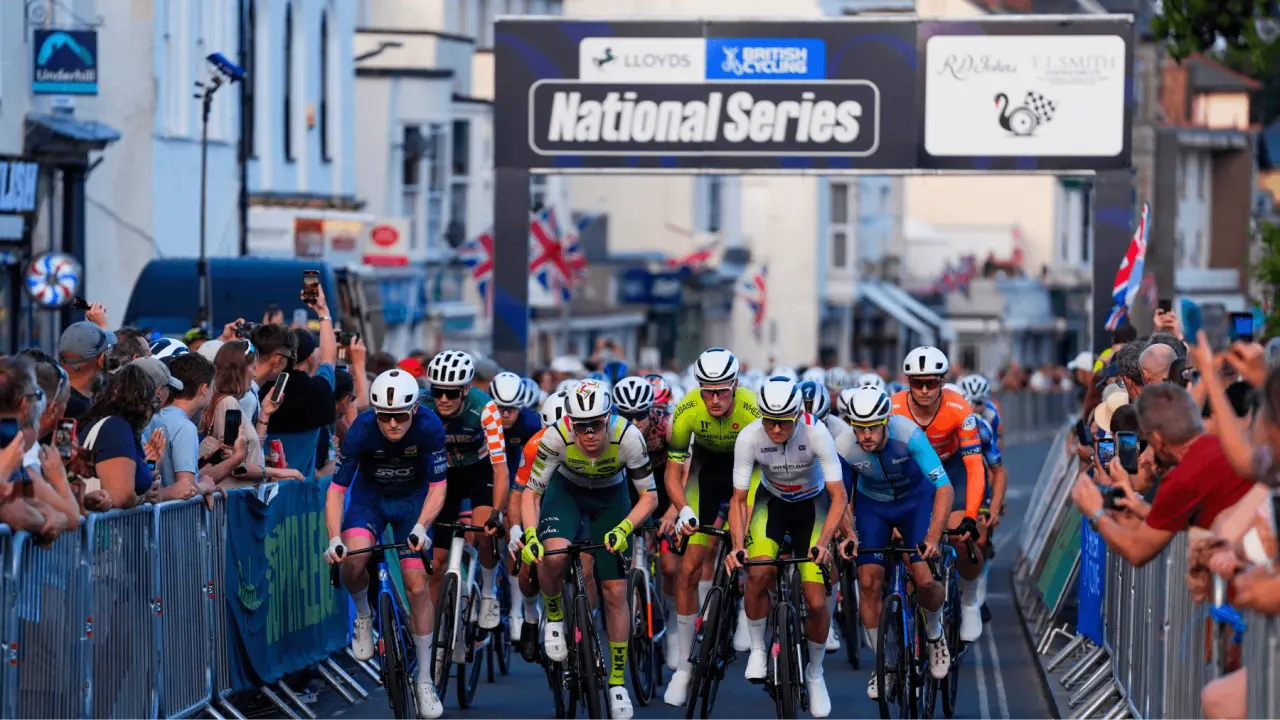 Professional cyclists riding through a city street under a National Series banner with spectators lining the barriers on both sides
