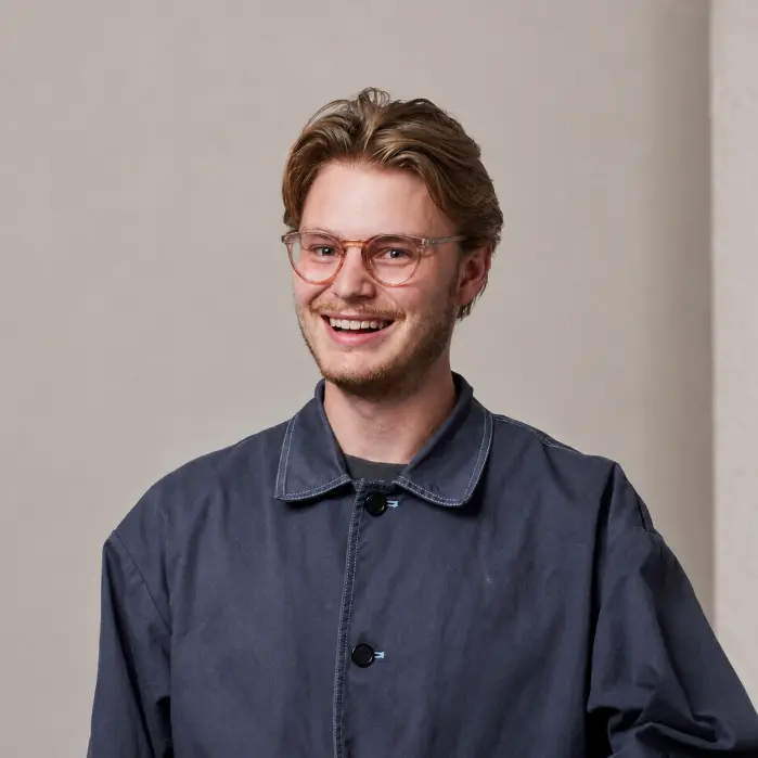 Man with brown hair and pink-rimmed glasses wearing a dark blue shirt, smiling at the camera against a neutral background