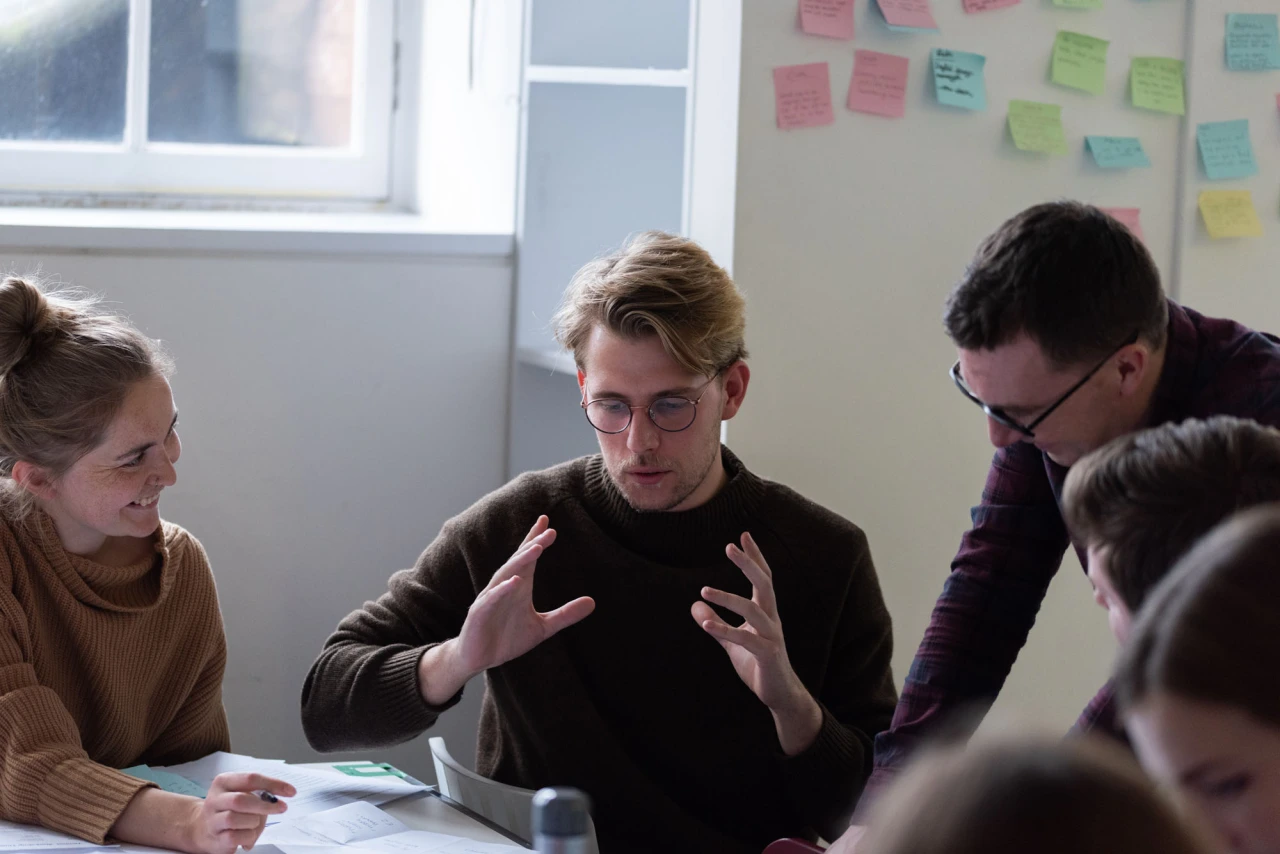Five people gathered around a table in a bright workshop space with coloured sticky notes on the wall behind them, discussing and collaborating on a project