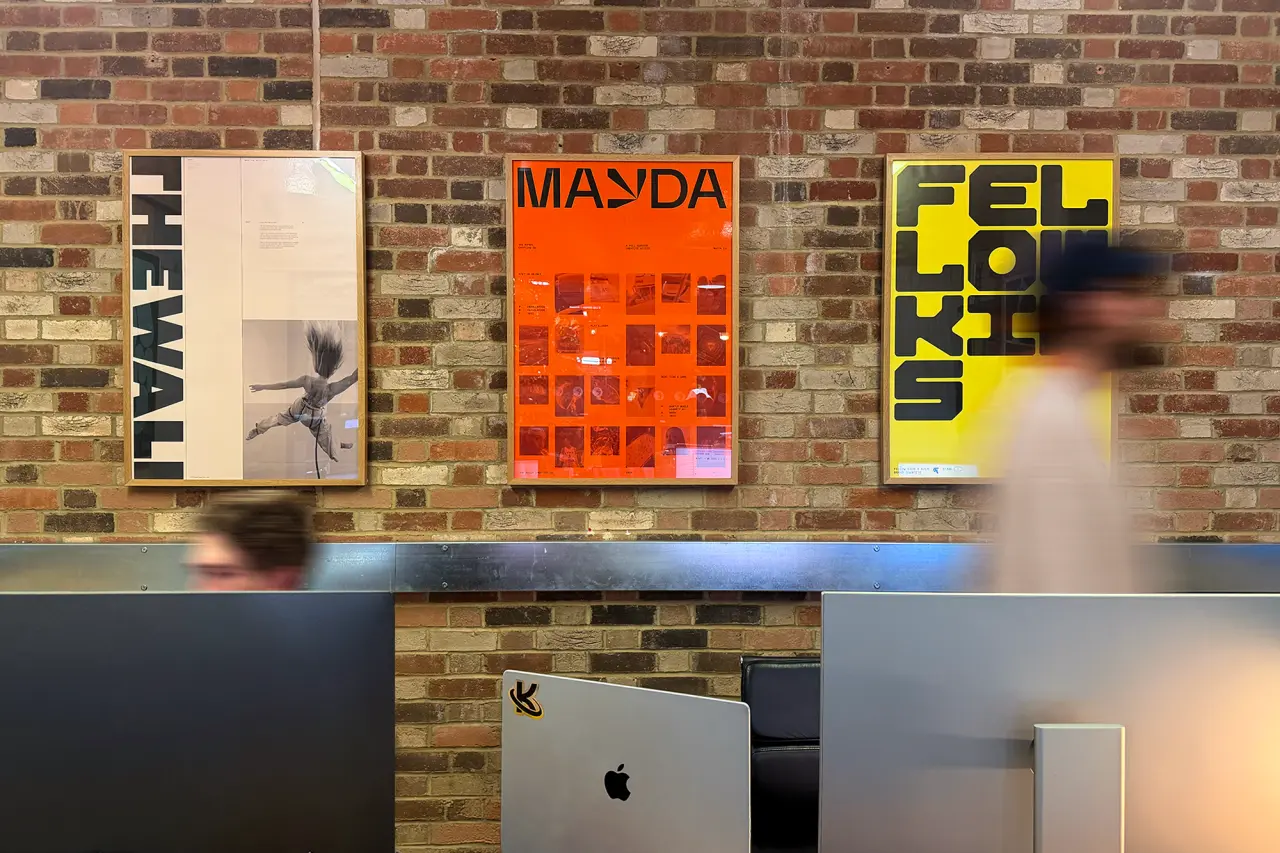 Three framed posters displayed on a brick wall above a desk, featuring Studio Kiln's bold typography and colour work in black, orange and yellow