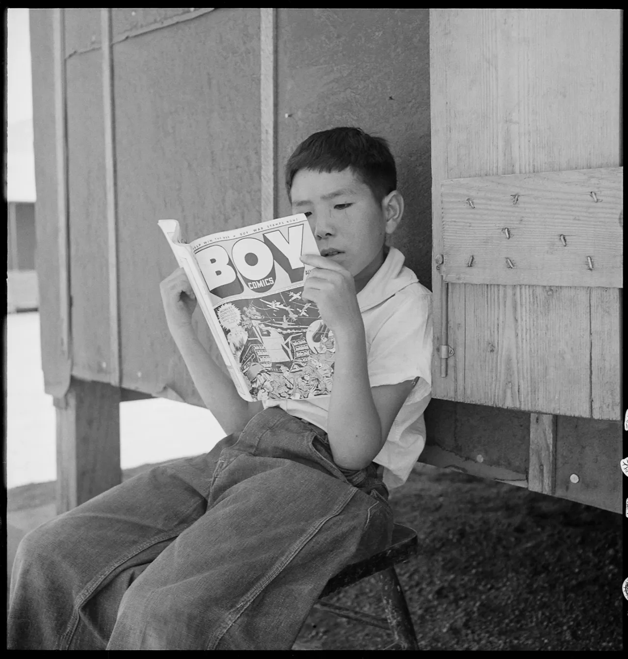 Young boy reading a comic book titled Boyz while seated against wooden barracks at Manzanar internment camp