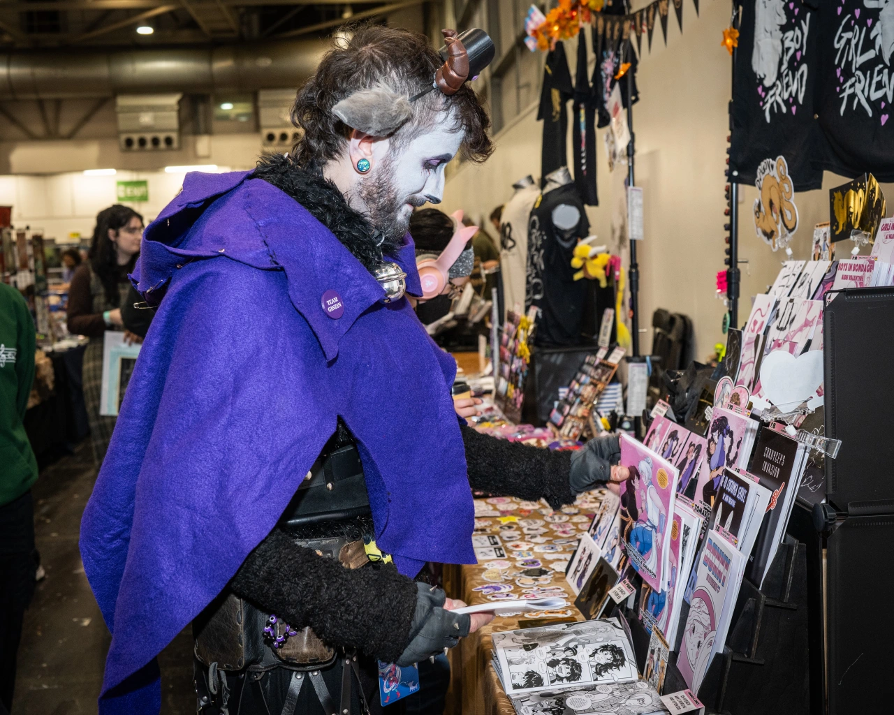 Person wearing purple coat and white face makeup examining zines and artwork displayed on a table at an indoor convention