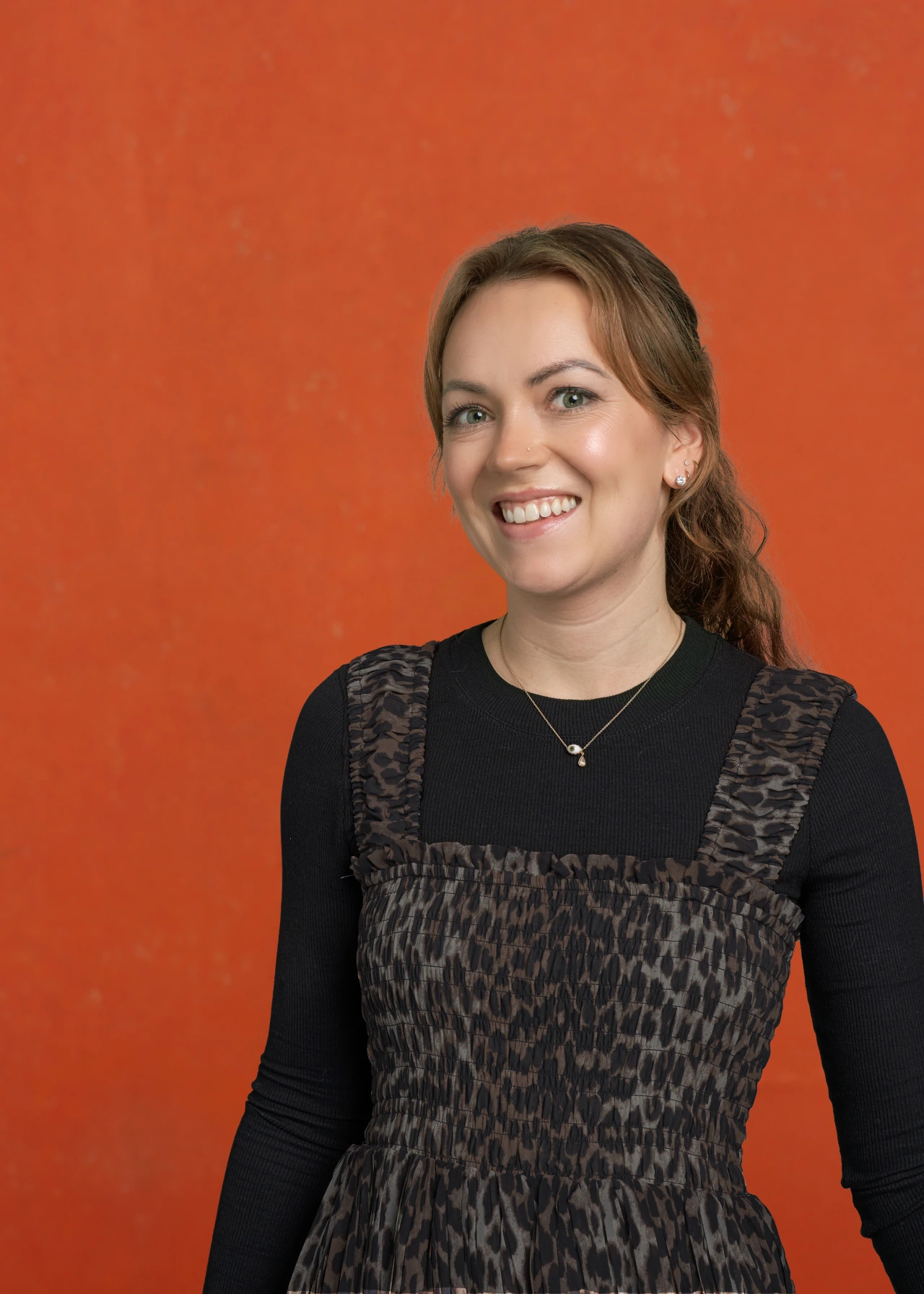 Merryn Mackinnon smiling against a bright orange background, wearing a black long-sleeved top and patterned sleeveless dress