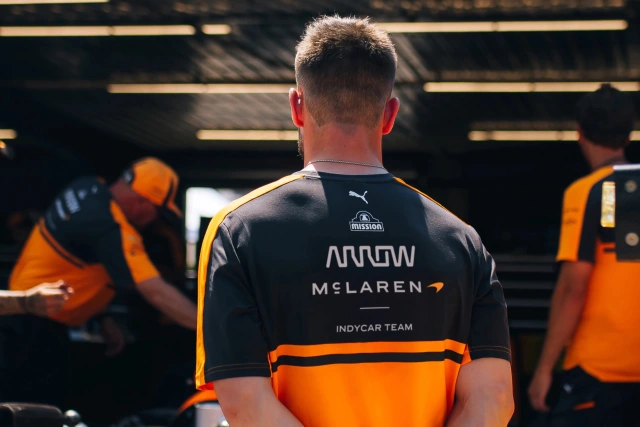 Member of McLaren IndyCar team wearing black and orange team kit with Arrow and McLaren wordmark visible on the back, standing in pit garage with teammates in background