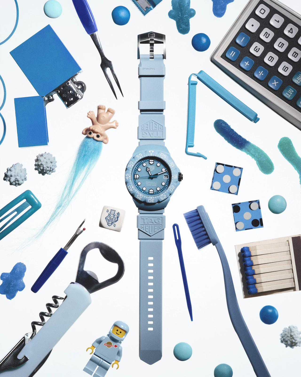 Flat lay arrangement of blue and grey objects including a light blue watch, calculator, pen, fork, toy figure, brushes, spheres and decorative items on white background