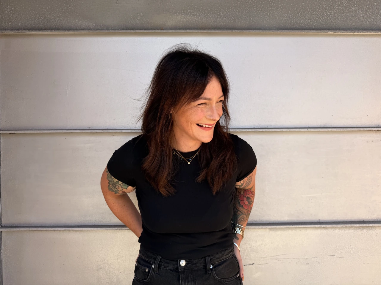 Cher Keane smiles whilst looking to the side, wearing a black t-shirt with visible tattoos on her arms against a white panelled wall