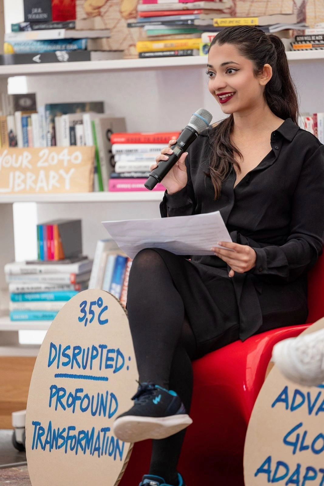 Woman in black shirt holding microphone and papers, speaking in front of a bookshelf with colourful books and motivational signs