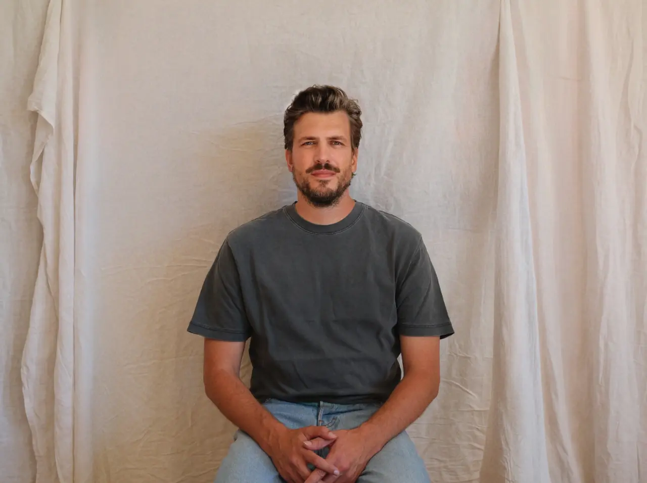 Jack De Caluwé. Man with dark hair and beard wearing a charcoal t-shirt and light blue jeans, standing against a cream curtained background