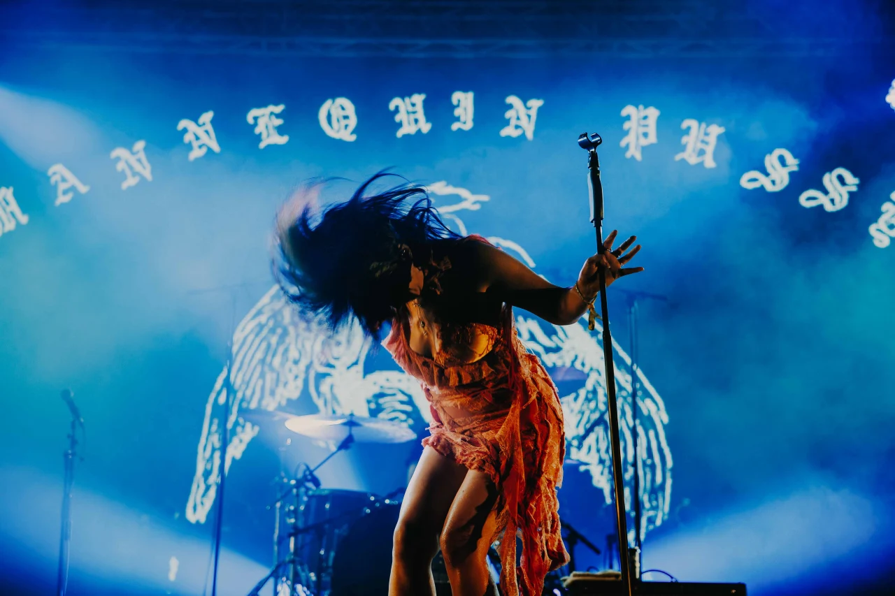 A singer in an orange lace dress flips her hair dramatically while performing onstage bathed in blue light and fog.